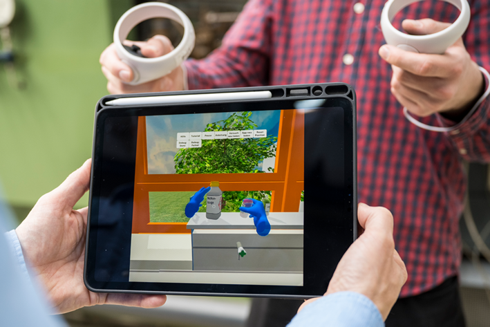 Ein Student steuert per Controller blaue Hände in einem virtuellen Labor. Der Vorgang im virtuellen Raum wird durch ein Tablet beobachtet.